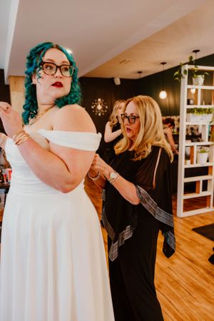 A bride in a white off-shoulder dress with teal hair getting ready in a room with pendant lights.