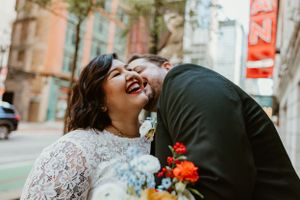 A joyful wedding couple embraces and laughs together on a city street with colorful florals.