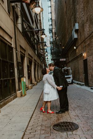 Couple shares intimate moment in urban alley with vintage lampposts and cobblestone street while bride wears red shoes.