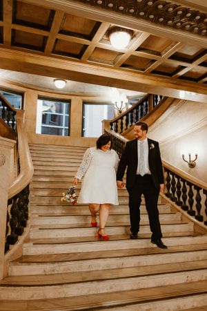 A couple descends a grand marble staircase with wooden coffered ceiling and romantic lighting.