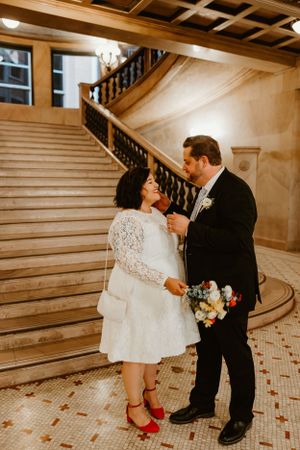 A romantic moment between newlyweds on an elegant marble staircase with the bride in red shoes and lace dress.