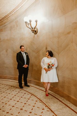 A couple poses in an elegant curved hallway with marble walls and decorative wall sconce lighting.