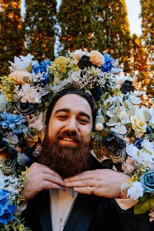 A bearded person wearing a black suit poses in front of autumn trees with a floral wreath made of blue and white flowers.