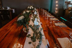 A rustic wooden reception table decorated with white linens, greenery garlands and glowing lights for a wedding celebration.
