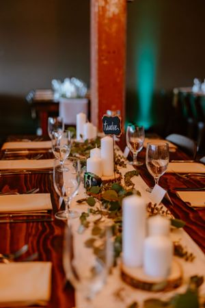 Elegant wedding reception table setting with white candles, greenery, and wine glasses on dark wooden tables.