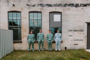 Wedding party stands in a row wearing matching sage green suits against a brick building.