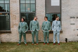 Groomsmen in coordinated mint colored suits pose together on grass by brick wall.