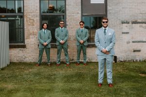 Groomsmen in matching sage green suits pose outside a brick building.