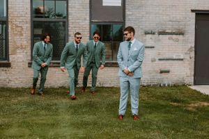 Group in coordinating sage and light blue suits pose casually on grass outside brick building.