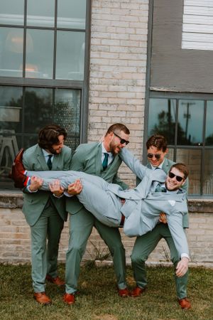 Groomsmen in matching sage green suits playfully lift and carry someone while posing outside a brick building.
