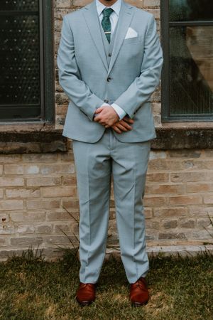 A groom in a light blue suit with green tie and brown shoes stands against a brick wall.
