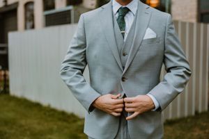 Close-up of a groom adjusting his light grey suit jacket with emerald tie at an outdoor wedding setting.