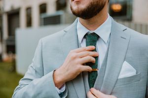 Closeup of a groom adjusting his dark tie against a light gray suit jacket with white pocket square.