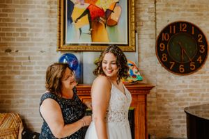Wedding dress fitting scene with detailed lace gown against brick wall interior with vintage clock.