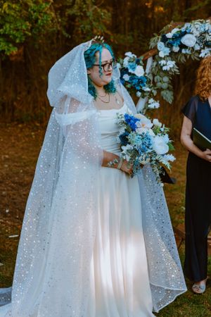 A bride in a sparkly white wedding gown with blue tinted hair holds a blue and white floral bouquet in a forest setting.