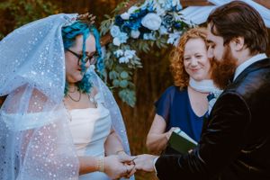 An intimate outdoor wedding ceremony under trees with a blue-themed floral arch.