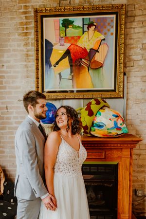 Colorful modern art painting in gold frame hangs on exposed brick wall above wooden fireplace mantel.