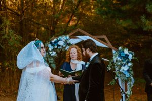 A wedding ceremony taking place under a floral arch adorned with blue and white flowers in a wooded setting at sunset.