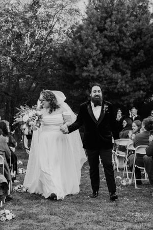 A joyful wedding couple walks down the aisle after their outdoor ceremony with guests seated in folding chairs.
