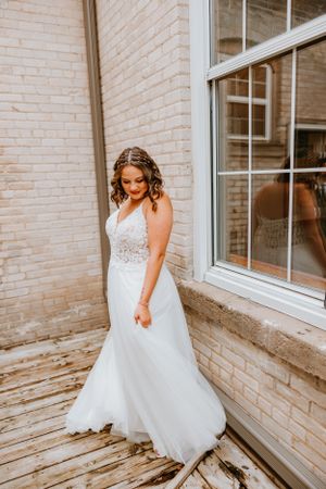 A bride in a flowing lace wedding gown poses beside a white brick wall and window.
