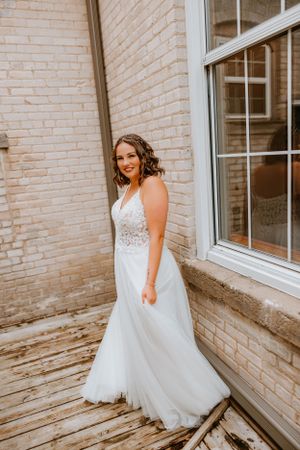 A bride in a flowing lace wedding gown stands against a cream brick wall and window.