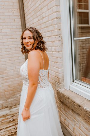 A lovely bride in a white lace wedding gown with spaghetti straps poses against a light brick wall on her special day.