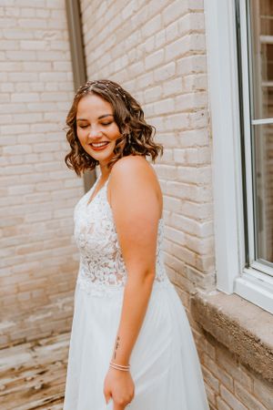 Person in white lace wedding dress smiles while standing against brick wall.