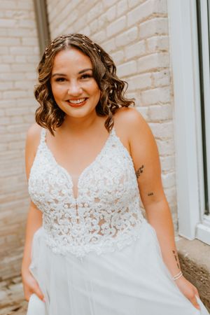 Bride in elegant lace wedding dress smiles radiantly against cream brick wall.