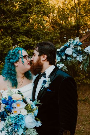 A joyful wedding couple share a tender moment outdoors, featuring bright blue floral bouquet and autumn foliage.