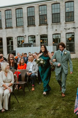 Wedding party members walk down aisle at outdoor ceremony by historic brick building with guests watching.