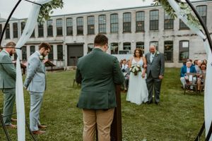 Outdoor wedding ceremony taking place under white arch with historic building in background.