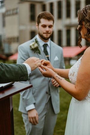 A wedding ceremony exchange of rings outdoors with the couple in elegant gray and white attire.
