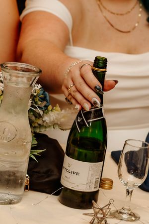 Close-up of hands opening a Westcliff wine bottle next to a glass carafe and wine glass on white tablecloth.