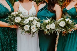 Wedding party holding elegant white rose and greenery bouquets with emerald green bridesmaid dresses.