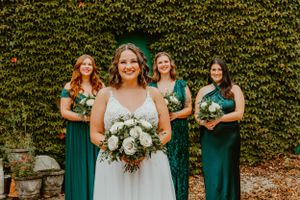 Wedding party poses outdoors with bridesmaids in emerald green dresses against ivy-covered wall.