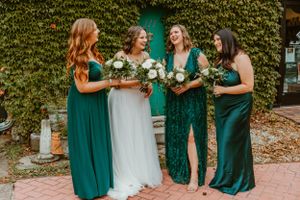 Bridal party laughs together while holding white floral bouquets against green ivy backdrop.