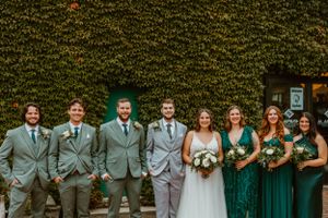 A wedding party poses outdoors against an ivy-covered wall wearing gray suits and emerald green dresses.