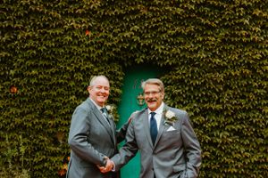 Two people in grey suits share a joyful moment in front of an ivy-covered wall with a green door.