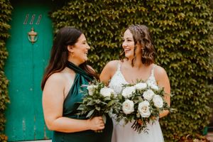 Two people sharing a joyful moment while holding white rose bouquets against an ivy-covered wall.