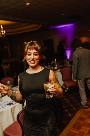 Person in a black sleeveless dress enjoying a drink at an indoor event with purple lighting and a dance floor.