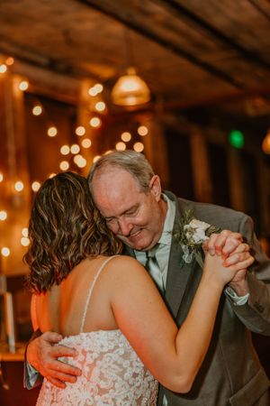 Father-daughter dance at wedding reception with warm string lights and rustic wooden beams overhead.