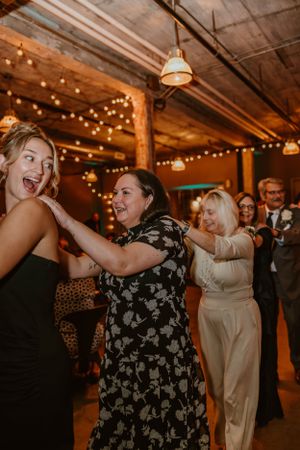 Wedding guests dance joyfully under string lights in an industrial venue with exposed wood beams and brick walls.