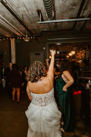 A bride in a white lace gown dances with her arm raised beneath industrial lighting and string lights at a reception.