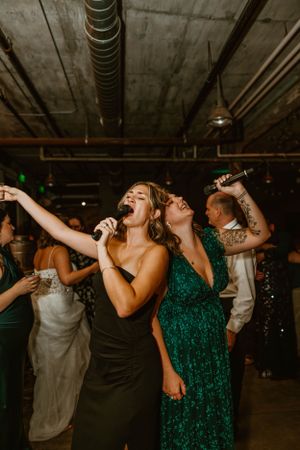 Wedding party guests singing karaoke and dancing enthusiastically in a dark industrial venue with exposed ceiling.