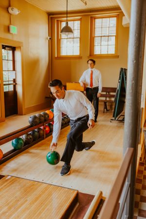Two people in formal attire bowling in a vintage-style bowling alley with wooden lanes.