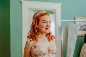 A smiling person in a floral wedding dress stands in front of a mint green wall wearing a delicate crystal headpiece.