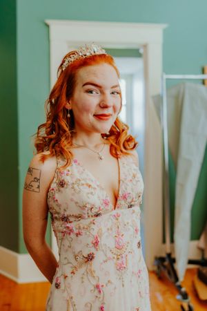A woman in a floral dress and tiara smiles while getting ready in a mint green room.