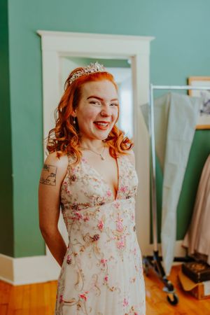 Person in a floral dress with a tiara smiling in front of a mint green wall.