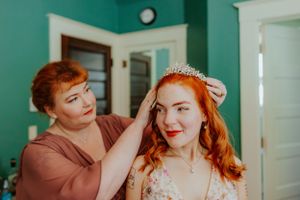 A bride with red hair wearing a tiara and wedding dress gets help with final preparations in a turquoise-walled room.