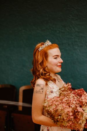 Elegant bride in crystal tiara holds pink hydrangea bouquet against dark green background.